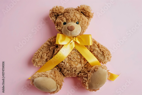 Top view of yellow awareness ribbon on teddy bear with bow on pink background, international photo