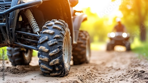 Close-up of an ATV tire on a dirt path, with a blurred rider in the background, illuminated by warm sunlight in a natural setting.