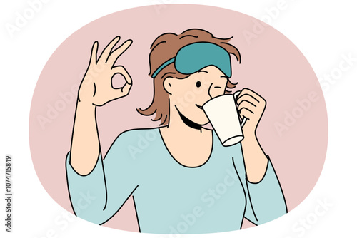 Woman drinks morning coffee after waking up and shows ok gesture standing in sleep mask and pajamas. Girl winks enjoying hot coffee or tea drink to cheer up and get ready for work day