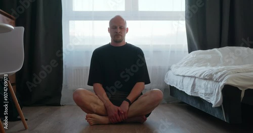 A bald man in a black T-shirt is doing breathing exercises sitting on the bedroom floor against the background of a window during quarantine. A man meditates in the morning during quarantine. 4k