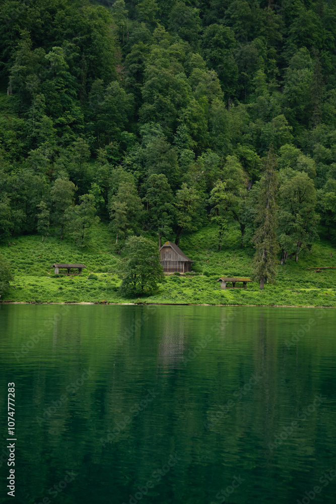 Obraz premium Secluded wooden cabin in lush forest along Königssee, Berchtesgaden National Park, Germany. The calm lake reflects vibrant greenery, creating a peaceful and harmonious natural scene.