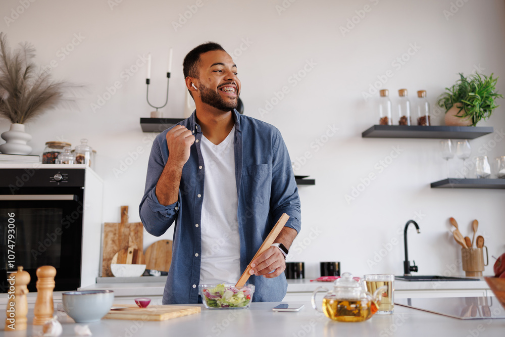 Cheerful african american man in earphones mixing fresh salad in kitchen at home Cheerful african american man in earphones mixing fresh salad in kitchen at home
