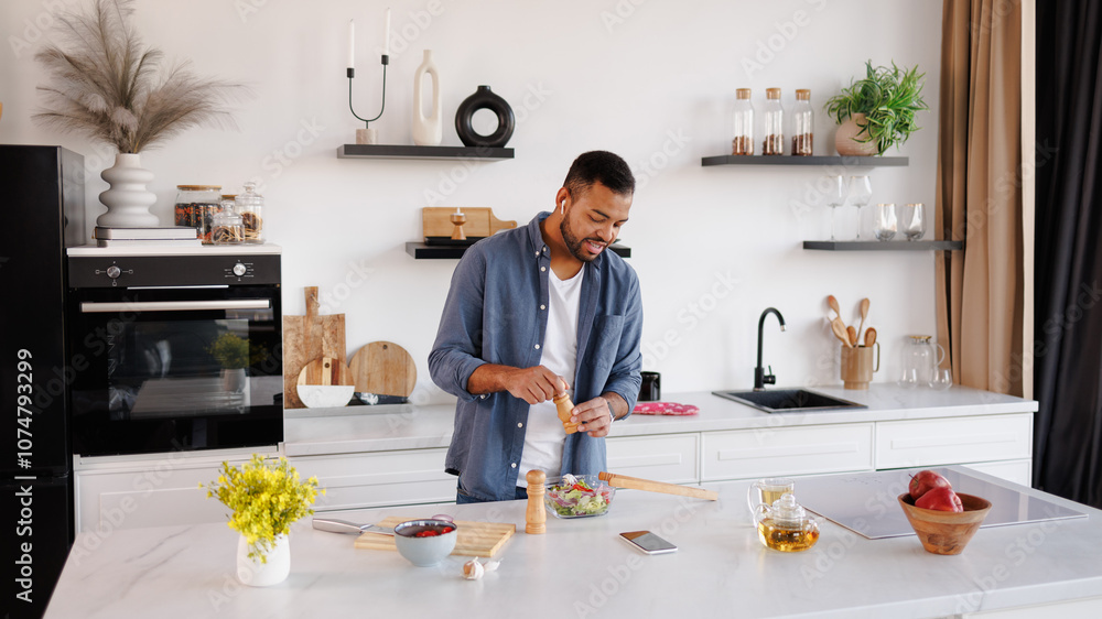 African american man in earphones seasoning fresh salad near tea and smartphone in kitchen African american man in earphones seasoning fresh salad near tea and smartphone in kitchen