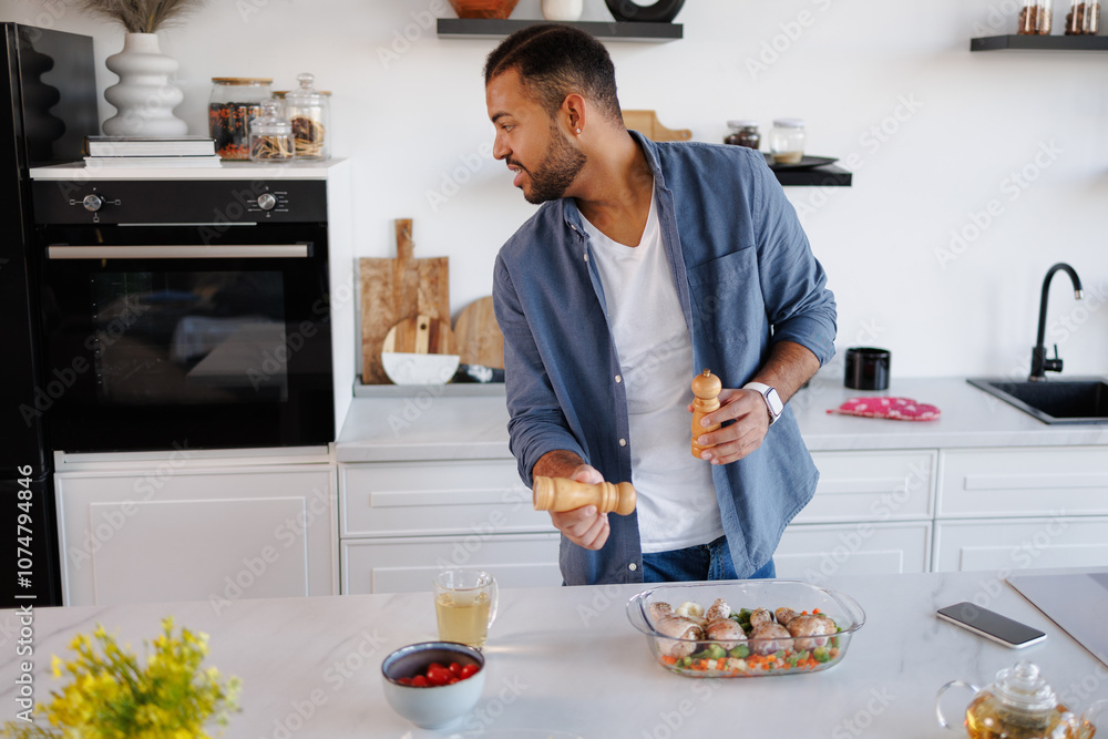African american man holding spices near food in baking dish and looking at oven in kitchen African american man holding spices near food in baking dish and looking at oven in kitchen