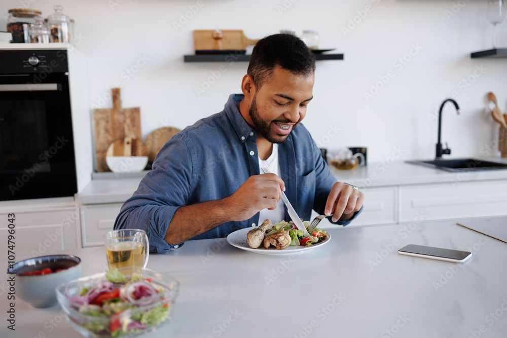 African american man smiling and eating fresh salad and chicken meat near tea and smartphone in kitchen African american man smiling and eating fresh salad and chicken meat near tea and smartphone in kitchen