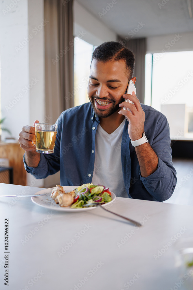 Smiling african american man talking on smartphone and holding herbal tea near healthy lunch on table at home Smiling african american man talking on smartphone and holding herbal tea near healthy lunch on table at home