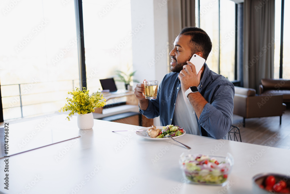 Smiling african american man talking on smartphone and holding cup of tea near lunch in kitchen Smiling african american man talking on smartphone and holding cup of tea near lunch in kitchen