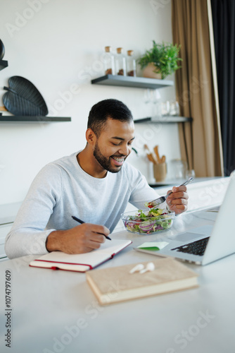 Smiling african american man eating fresh salad near laptop and writing on notebook in kitchen