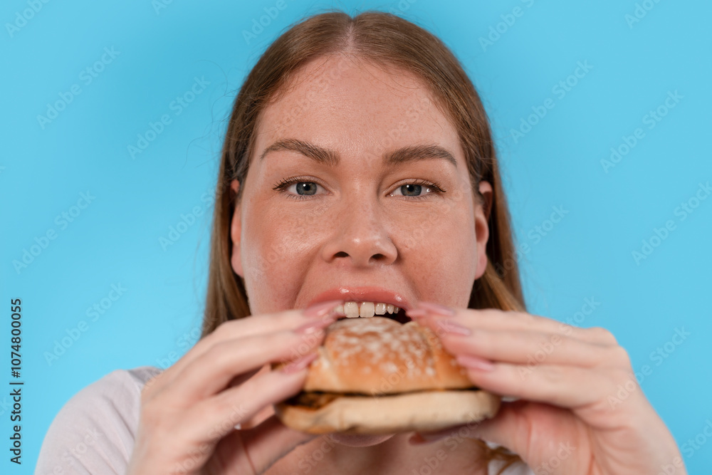 A woman eagerly prepares to take a big bite of a delicious hamburger against a bright blue background A woman eagerly prepares to take a big bite of a delicious hamburger against a bright blue background