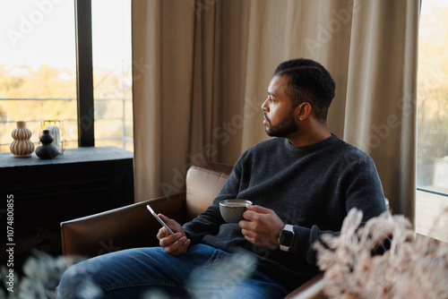 African american man holding coffee and smartphone while sitting on armchair at home