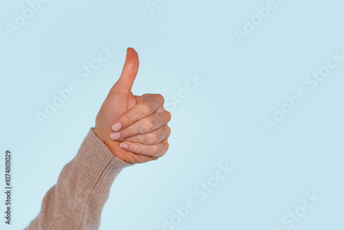 A hand making a thumbs-up gesture against a light blue background, expressing approval or positivity in a casual setting photo