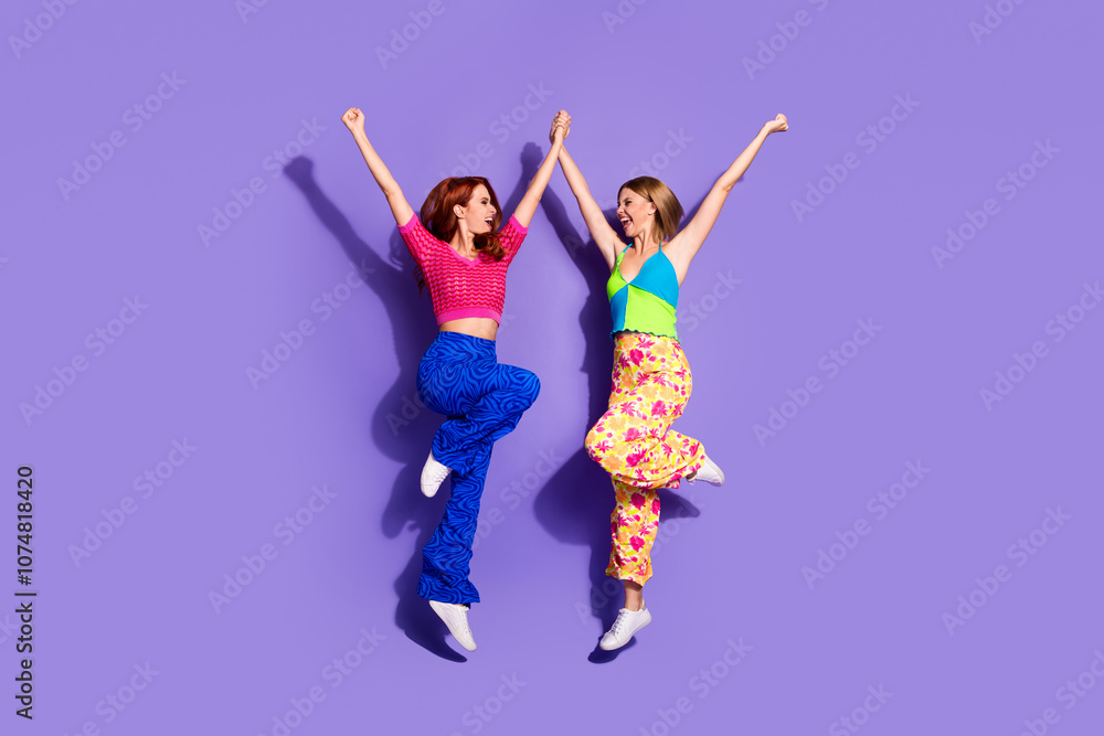 Full size photo of two pretty young girls hold hands winning jump wear trendy vintage outfit isolated on purple color background Full size photo of two pretty young girls hold hands winning jump wear trendy vintage outfit isolated on purple color background