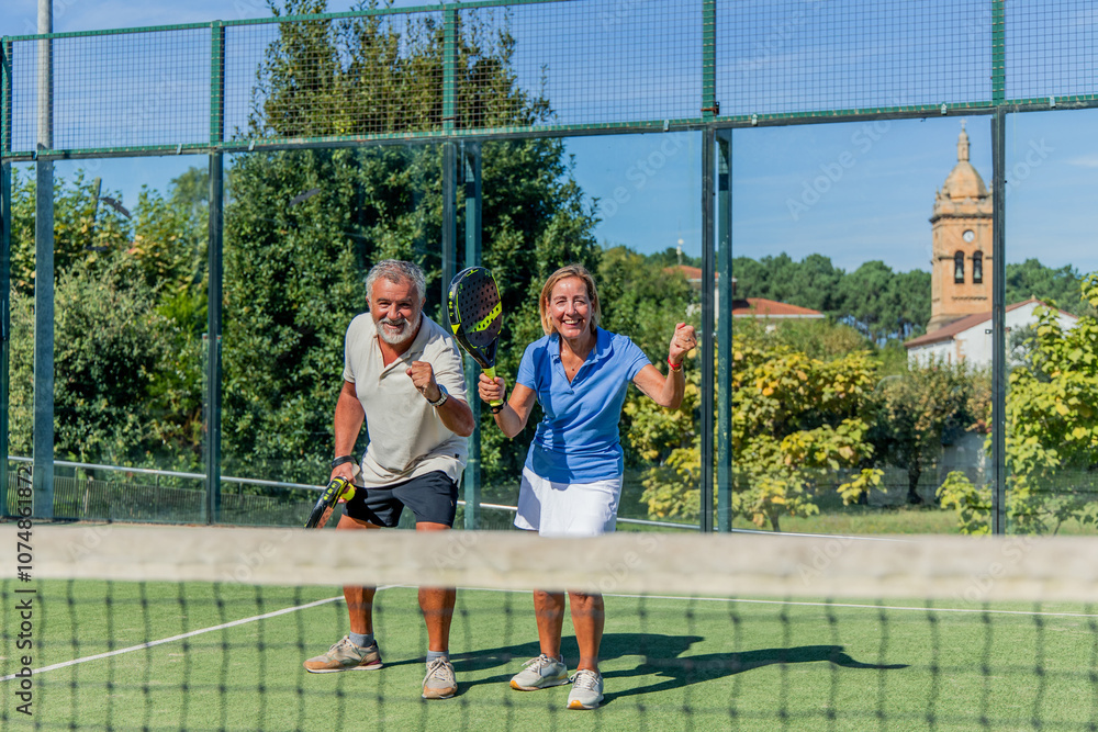 Senior elderly padel player couple celebrating victory on court with raised fists Senior elderly padel player couple celebrating victory on court with raised fists