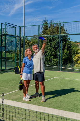 vertical elderly padel couple capturing post-match selfie on court with smartphone