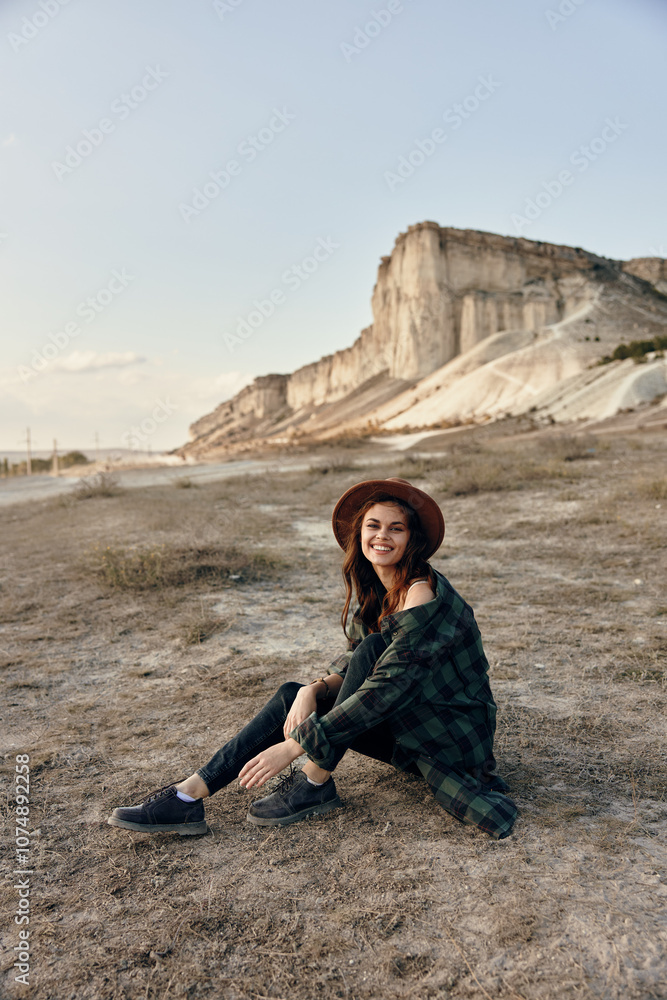 Naklejka premium Woman in a stylish hat sitting peacefully in front of a majestic cliff on a sunny day