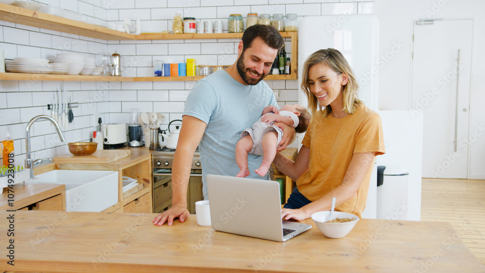Busy Family In Kitchen At Breakfast With Parents Working On Laptop And Caring For Baby Son Busy Family In Kitchen At Breakfast With Parents Working On Laptop And Caring For Baby Son