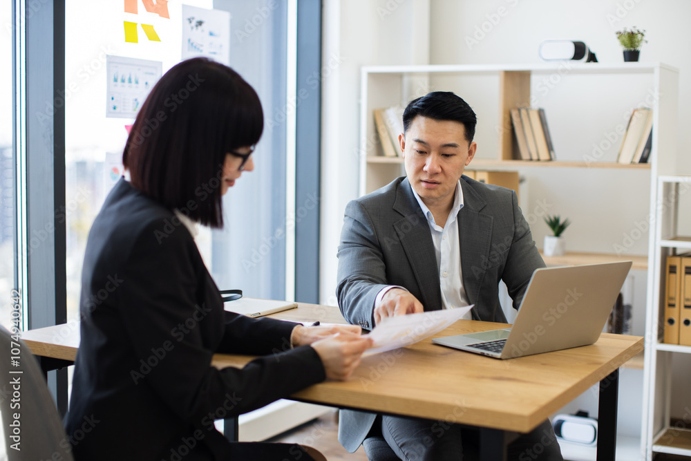 Asian businessman in grey suit reviews report with Caucasian businesswoman in a modern office. They are engaged in serious discussion, emphasizing teamwork and corporate environment. Asian businessman in grey suit reviews report with Caucasian businesswoman in a modern office. They are engaged in serious discussion, emphasizing teamwork and corporate environment.