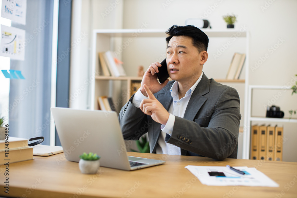 Asian male business professional mid-30s engaging in phone call while working on laptop in modern office setting. Depicts focused work environment with digital charts and documents. Asian male business professional mid-30s engaging in phone call while working on laptop in modern office setting. Depicts focused work environment with digital charts and documents.