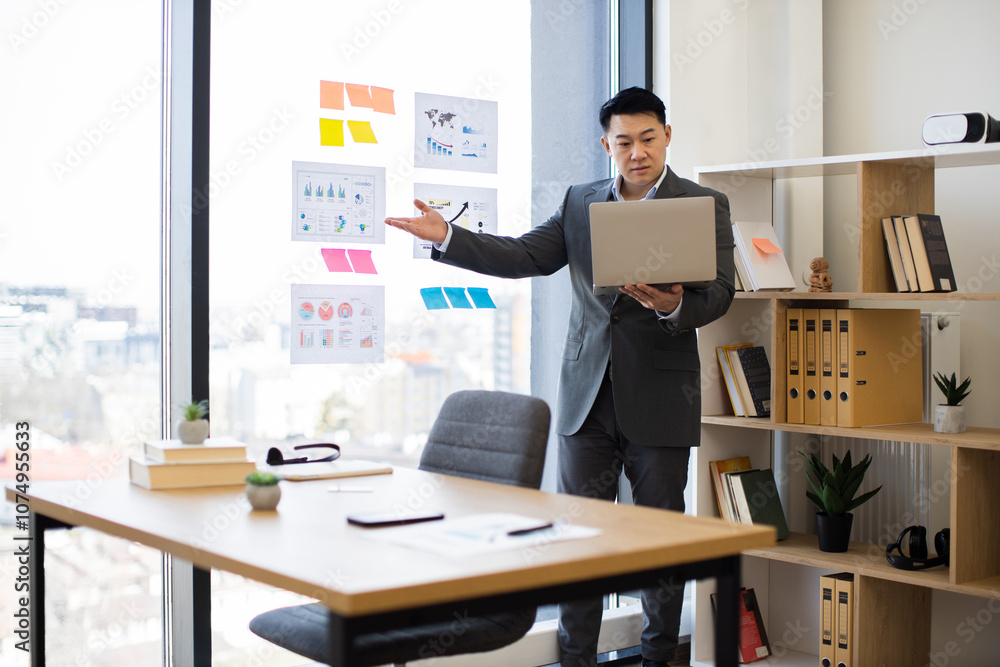 Asian businessman presents data charts on laptop during online video call. He stands in office near panoramic window, analyzing business graphs. Asian businessman presents data charts on laptop during online video call. He stands in office near panoramic window, analyzing business graphs.