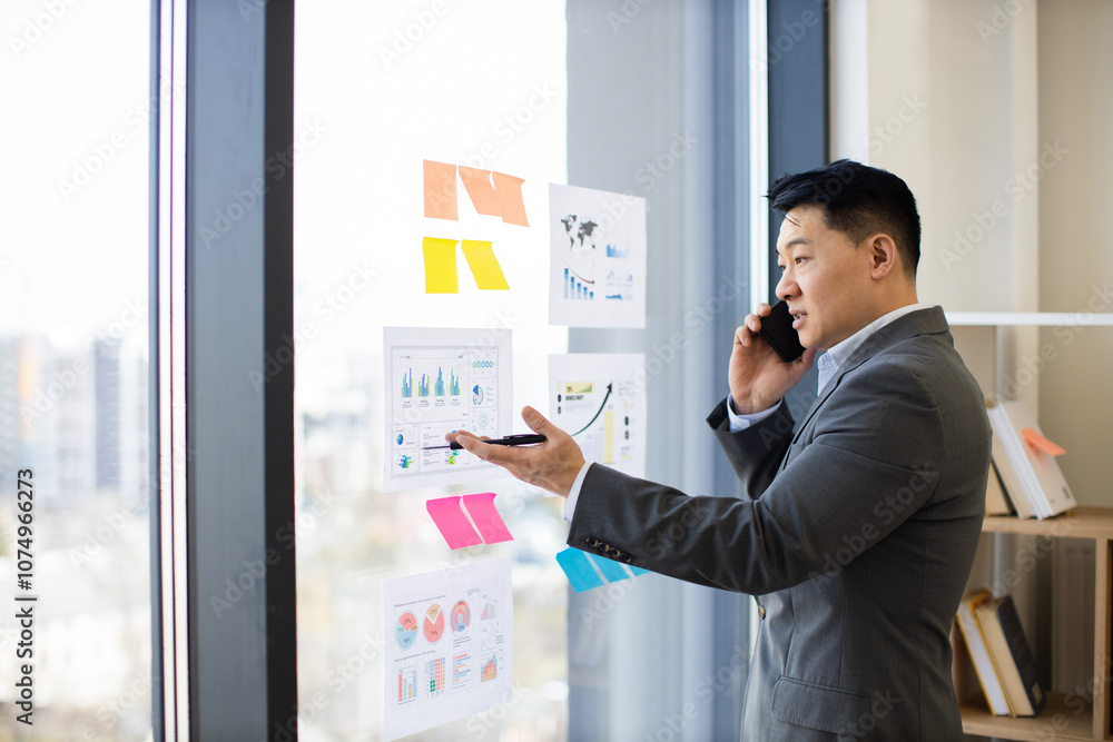 Asian male businessman stands at panoramic window, analyzing graphs and charts while on smartphone. Engaged in a professional conversation with a colleague, he exhibits focus and concentration. Asian male businessman stands at panoramic window, analyzing graphs and charts while on smartphone. Engaged in a professional conversation with a colleague, he exhibits focus and concentration.