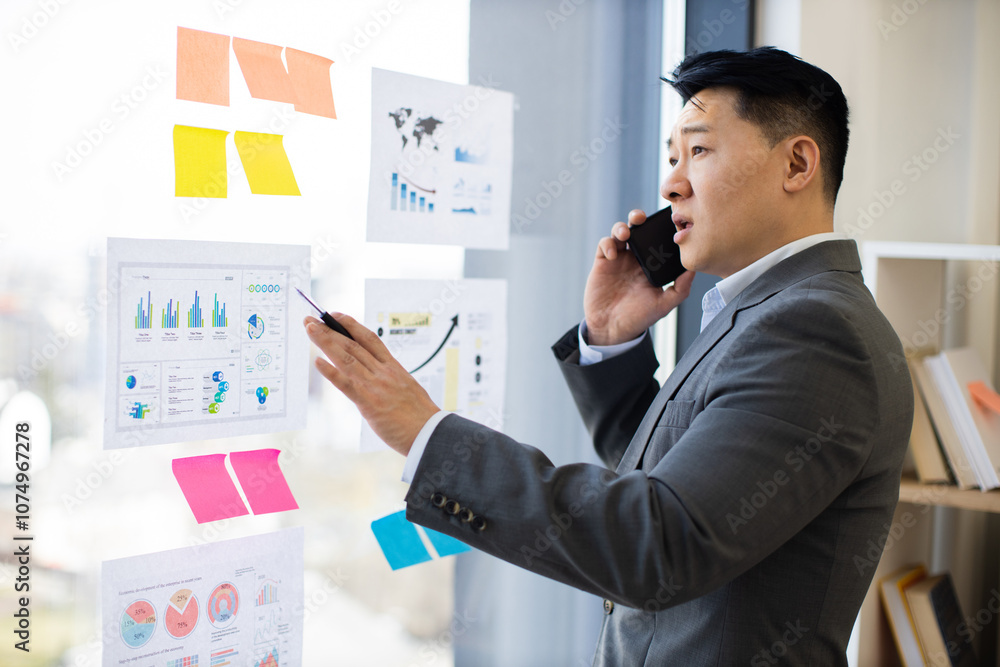 Asian male businessman in office setting, analyzing graphs and charts on panoramic window. He is calling colleague using smartphone for data insights. Asian male businessman in office setting, analyzing graphs and charts on panoramic window. He is calling colleague using smartphone for data insights.