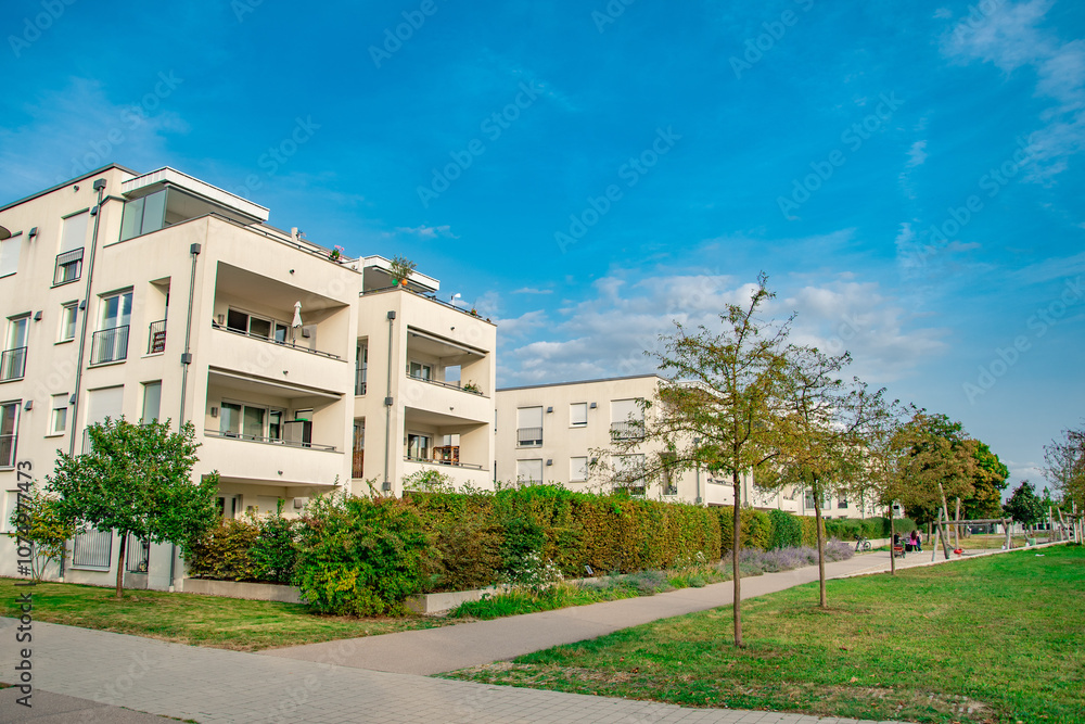 Modern apartment building surrounded by greenery under a clear blue sky. The residential complex features contemporary architecture with wooden accents, large windows, and balconies adorned with plant Modern apartment building surrounded by greenery under a clear blue sky. The residential complex features contemporary architecture with wooden accents, large windows, and balconies adorned with plant