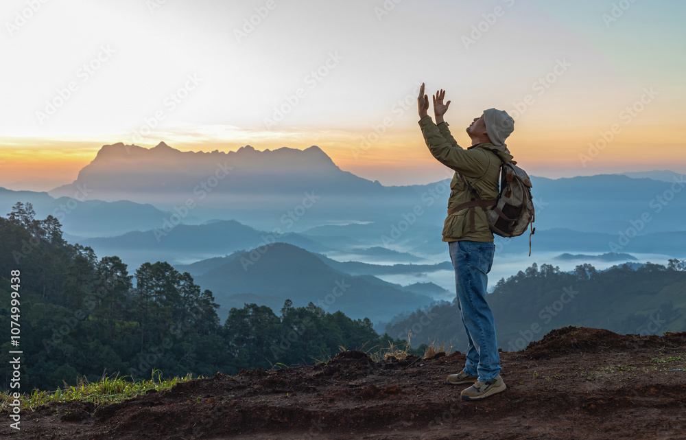 Young male hiker with backpack open palm up praying to God on the mountain sunset background Young male hiker with backpack open palm up praying to God on the mountain sunset background