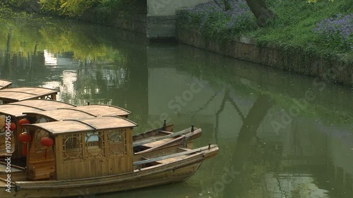 A peaceful video of traditional wooden boats docked along a serene canal in a lush, green garden. Red lanterns adorn the boats, creating a classic Eastern ambiance.