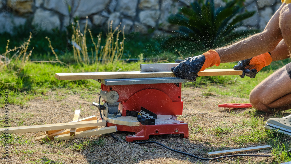 Fototapeta premium A middle-aged man cutting timber with a circular saw in the garden of his house on a summer day