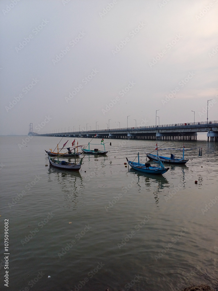 Busy Traffic on Suramadu Bridge in the Distance, with Floating Fisherman’s Boats in the Foreground, Highlighting the Contrast Between Modern Infrastructure and Traditional Maritime Life Busy Traffic on Suramadu Bridge in the Distance, with Floating Fisherman’s Boats in the Foreground, Highlighting the Contrast Between Modern Infrastructure and Traditional Maritime Life