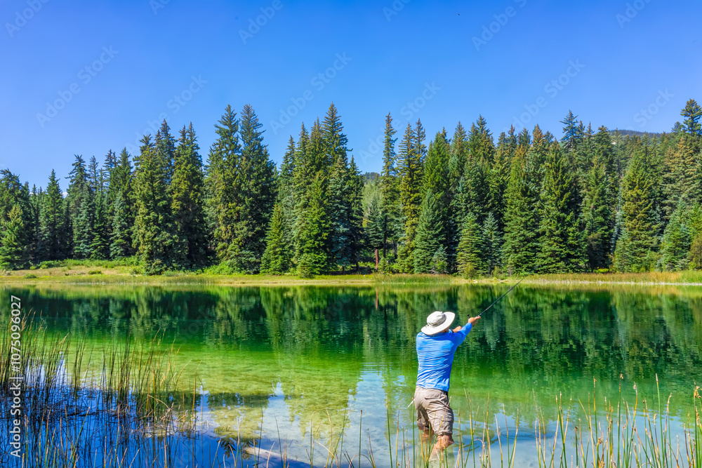 Eagle County Emerald Lake with coniferous trees and mirror sky reflection on water surface and man fishing with rod and reel inside Eagle County Emerald Lake with coniferous trees and mirror sky reflection on water surface and man fishing with rod and reel inside