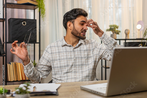 Overworked young Indian bearded businessman suffering from eye strain and headache working on laptop sitting at home office desk. Tired freelancer takes off glasses and massages, rubs bridge of nose. photo