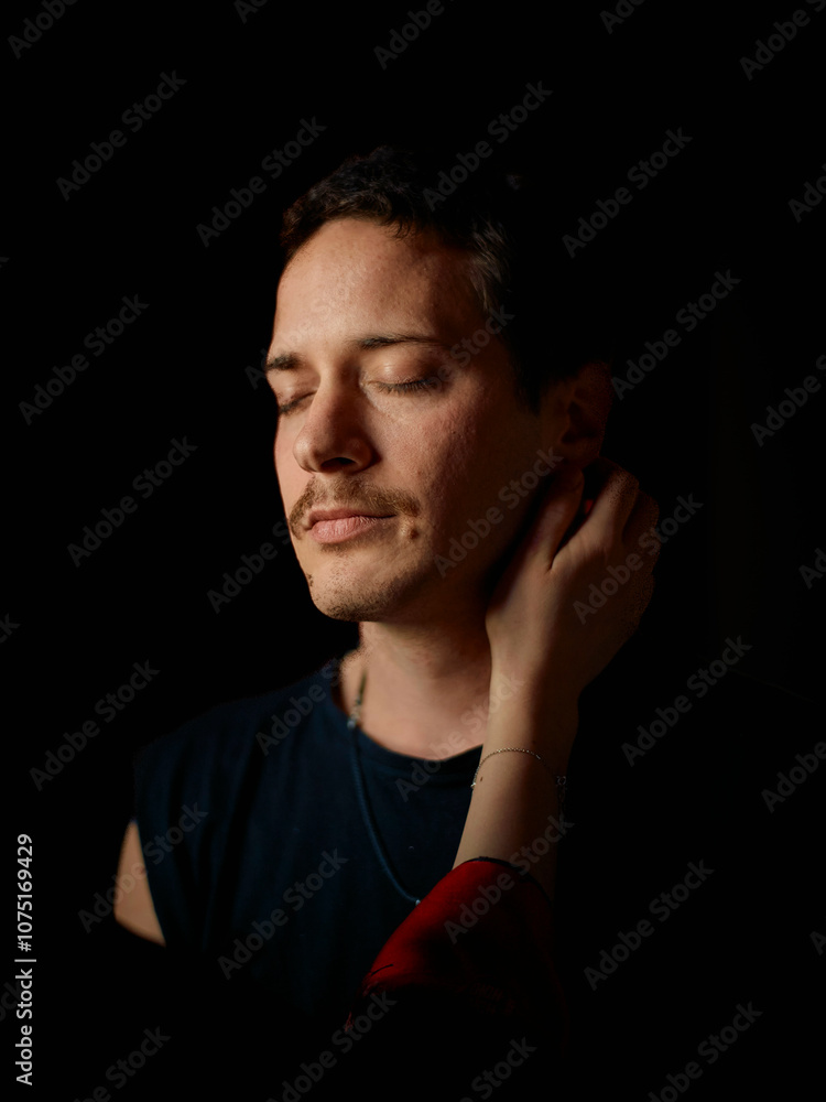 A contemplative man with closed eyes stands in a dark background, his face partially illuminated, while his partner's hand gently touches the side of his neck. A contemplative man with closed eyes stands in a dark background, his face partially illuminated, while his partner's hand gently touches the side of his neck.