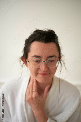 Close-up of a woman with glasses and a thoughtful expression wearing a white robe touching her neck gently against a neutral background. photo