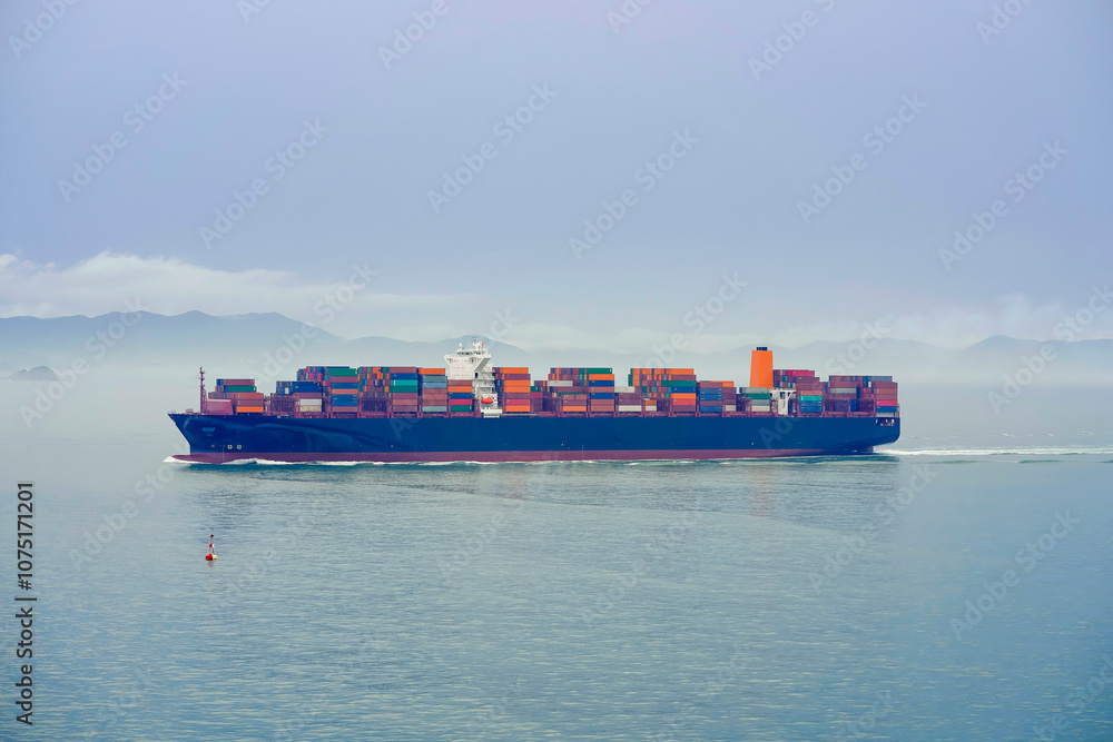 A cargo ship laden with containers sails across a misty sea with mountains faintly visible in the background, Busan, Korea A cargo ship laden with containers sails across a misty sea with mountains faintly visible in the background, Busan, Korea