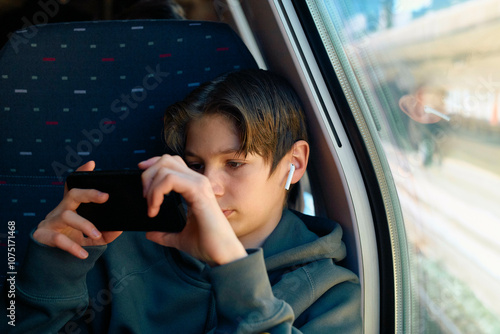 A teenager wearing a hoodie and wireless earbuds focuses intently on his smartphone while sitting by a train window. photo