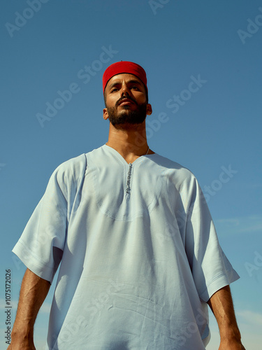 Confident man standing against a clear blue sky wearing a white tunic and red headband, Morocco