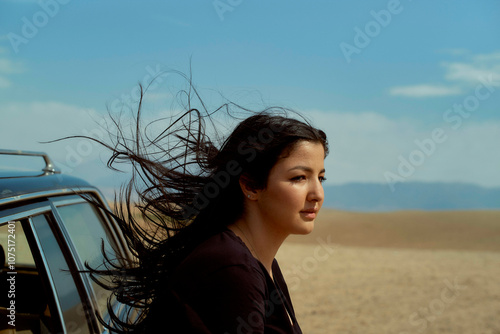 Woman with flowing hair looking into the distance next to her car in a desert setting, Morocco photo