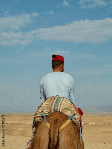 A man wearing a red headband and white shirt rides a camel in the desert under a clear blue sky, Morocco photo