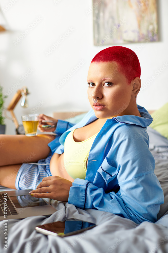 Fototapeta premium A young woman relaxes at home, sipping tea and using her laptop in comfortable clothing.