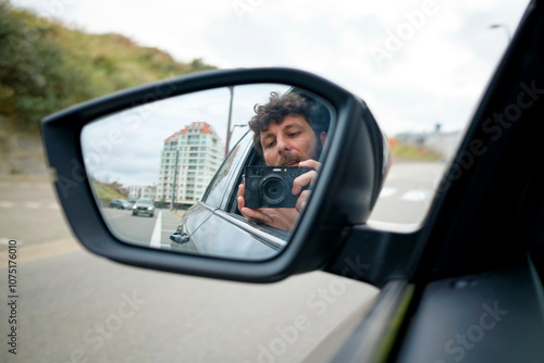 A man takes a photo of himself in the side mirror of a car, with buildings visible in the background, Belgium photo