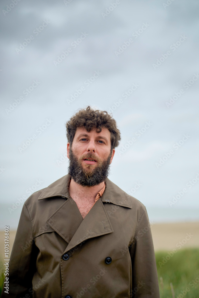 A man with a beard wearing a trench coat stands in front of a cloudy sky and beach, Belgium A man with a beard wearing a trench coat stands in front of a cloudy sky and beach, Belgium