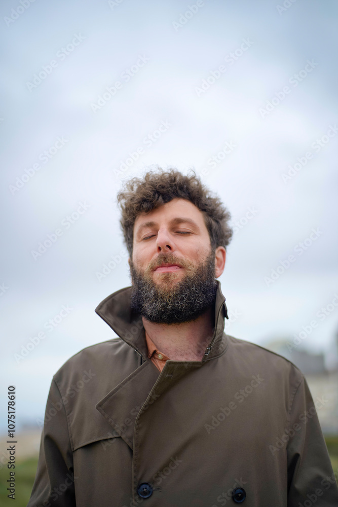 Man with curly hair and beard enjoying the fresh air with eyes closed and a content expression, wearing a brown coat, Belgium Man with curly hair and beard enjoying the fresh air with eyes closed and a content expression, wearing a brown coat, Belgium