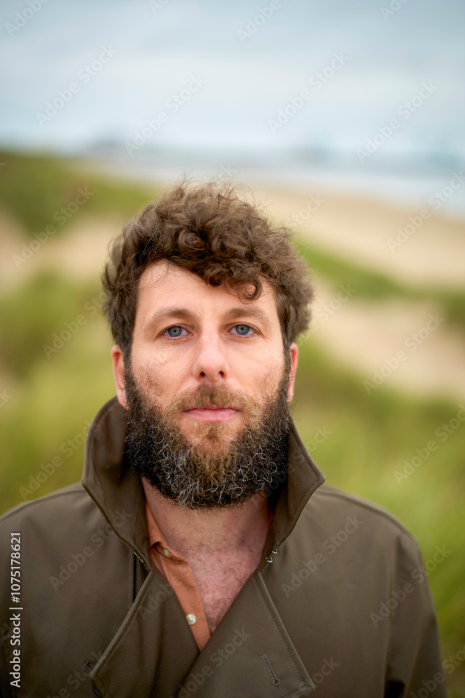 Man with curly hair and a beard wearing a green jacket standing against a beach dunes background, Belgium Man with curly hair and a beard wearing a green jacket standing against a beach dunes background, Belgium