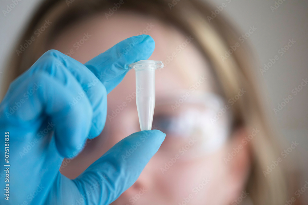 A close-up view of a gloved hand holding a clear test tube in front of a female scientist wearing protective goggles. A close-up view of a gloved hand holding a clear test tube in front of a female scientist wearing protective goggles.