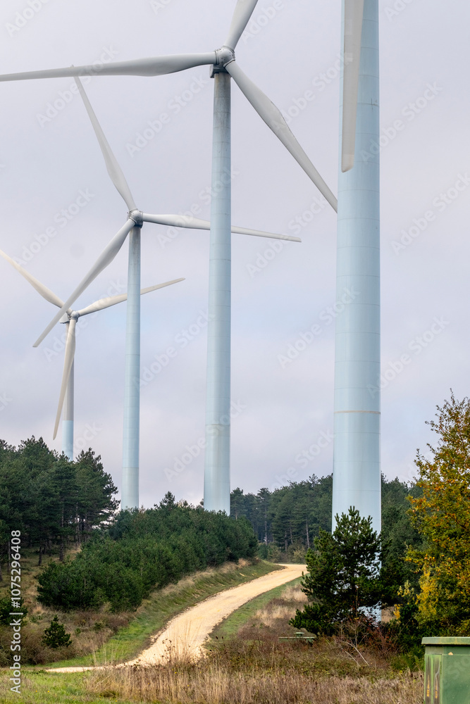 A close-up perspective showcasing several tall wind turbines, positioned along a dirt path, surrounded by greenery and trees, under a cloudy sky in a wide open space in Navarra Spain A close-up perspective showcasing several tall wind turbines, positioned along a dirt path, surrounded by greenery and trees, under a cloudy sky in a wide open space in Navarra Spain