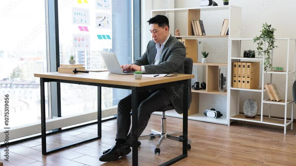 Asian businessman in modern office working on laptop. Mid-adult male in professional attire focused on work. Bright office space with large window, organized workspace, and stylish decor. Asian businessman in modern office working on laptop. Mid-adult male in professional attire focused on work. Bright office space with large window, organized workspace, and stylish decor.