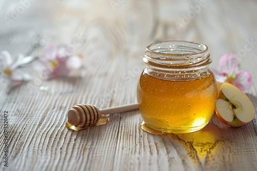 A jar of honey with a wooden dipper, apple slices, and pink flowers on a wooden surface.