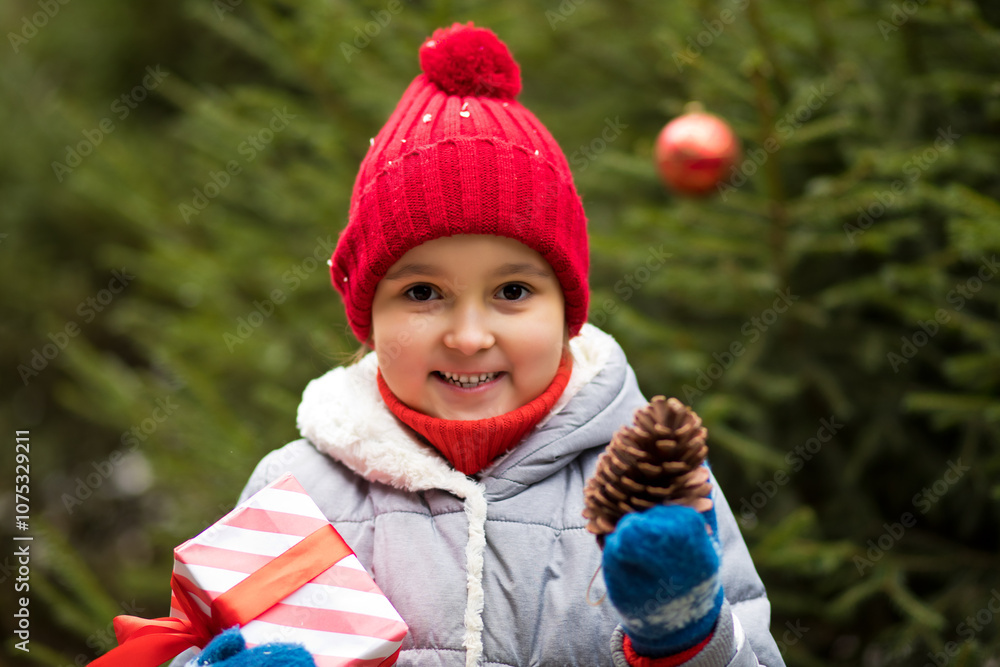 Happy little smiling girl in red cap with Christmas gift box standing near by spruce. Holiday celebrate concept. Happy little smiling girl in red cap with Christmas gift box standing near by spruce. Holiday celebrate concept.