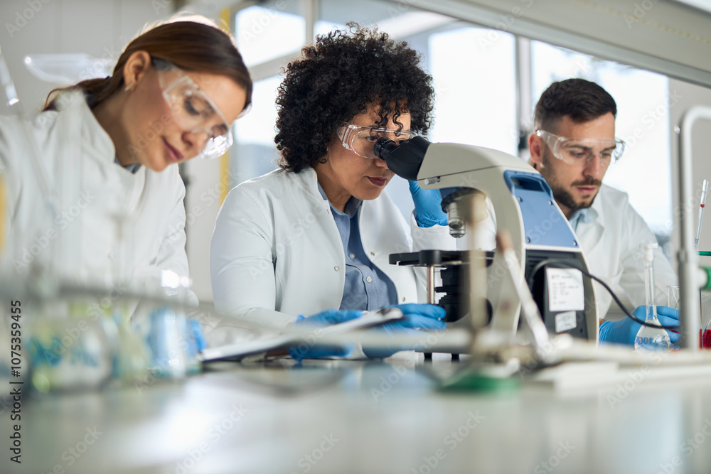 Obraz premium Mid adult female scientist looking through a microscope while working in laboratory with her colleagues.